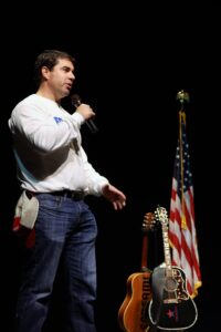A man holding a microphone and standing next to an american flag.