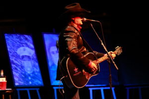 A man in black jacket holding a guitar on stage.