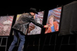 A man in black jacket standing next to paintings.